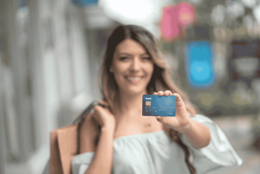 Jeune femme souriante tenant une carte bancaire devant elle dans un environnement urbain