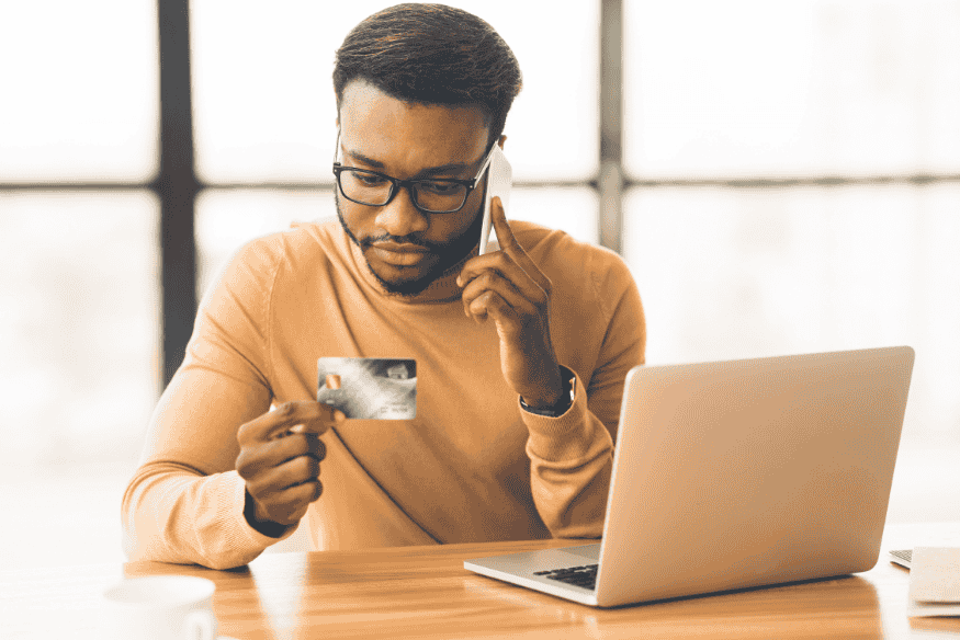 Uomo al telefono con carta bancaria in mano davanti al laptop, immagine che rappresenta le démarches utili per contestare un pagamento con carta bancaria.
