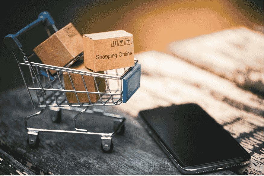 Un mini chariot de courses avec des cartons portant la mention “Shopping Online”, à côté d’un smartphone.