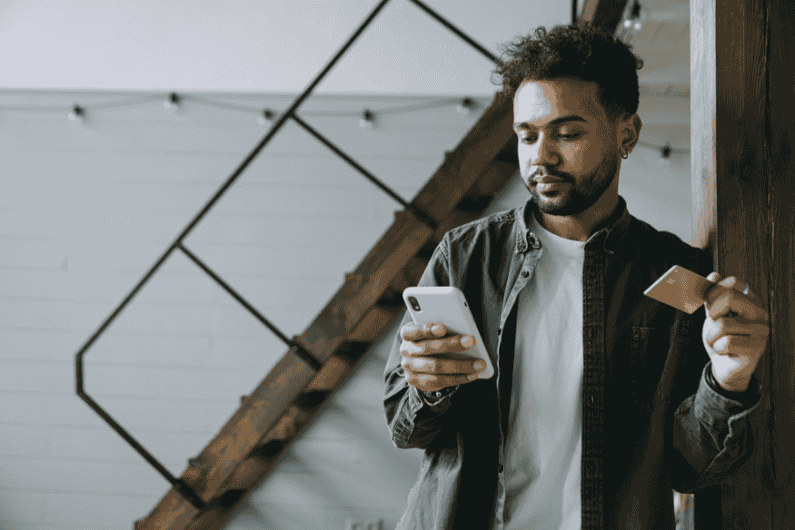 Uomo che controlla lo smartphone mentre tiene una carta bancaria in mano, immagine che rappresenta un pagamento rifiutato con carta bancaria e la verifica del conto.