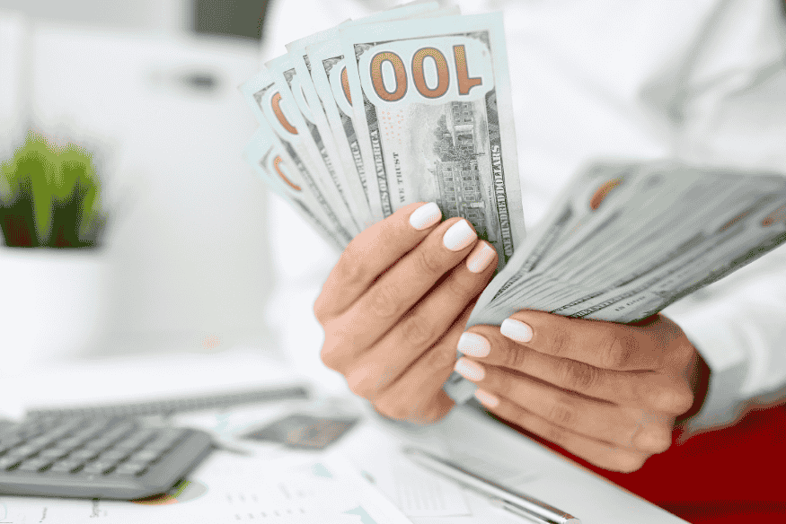 Femme comptant plusieurs billets de 100 dollars à son bureau, avec des documents financiers en arrière-plan.