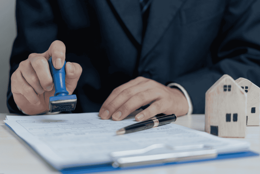 Main d’un professionnel tamponnant un contrat de prêt bancaire avec des maisons miniatures à côté.